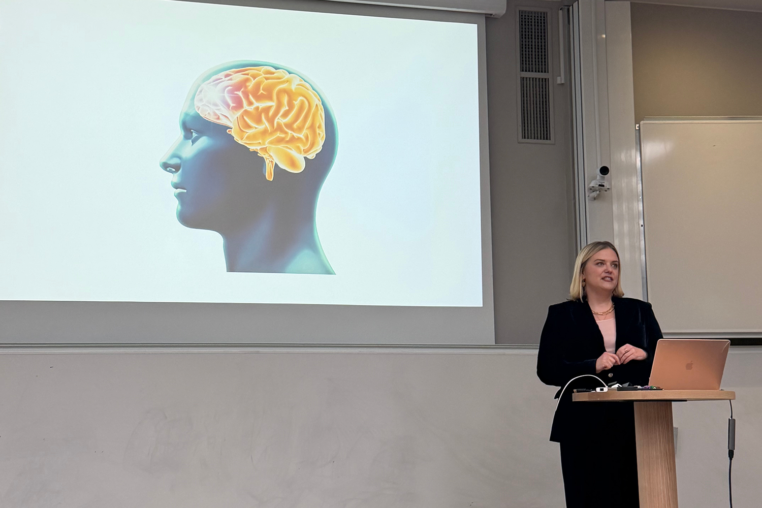 Adrienne speaking at a lectern with a presentation behind her showing a 3D generated image of a brain inside a human's head.