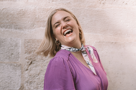 Adrienne in a light pinkish-purple blouse and light colored scarf tied around her neck. She's got her eyes closed as she tosses her short blonde bob, smiling enthusiastically.