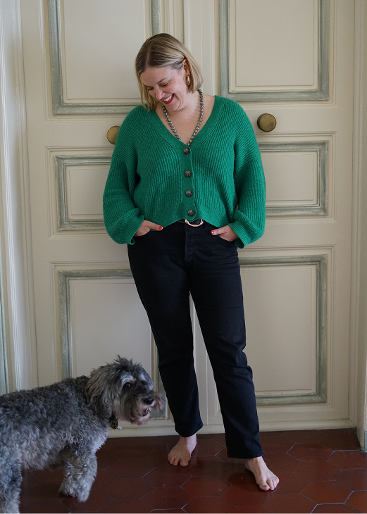 Adrienne in a baggy cropped green sweater, black pants, and bare feet, standing in front of a white double door with sage green trim. She's looking down at her medium-sized, curly-haired, black and gray dog.