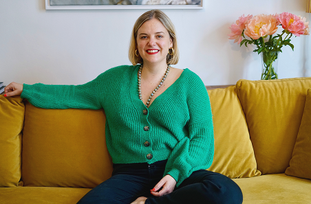 Adrienne in a baggy, cropped green sweater on a bright yellow couch.